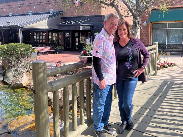 Smiling couple posing together on a small wooden bridge over a stream in a shopping and dining plaza with brick buildings and flower planters